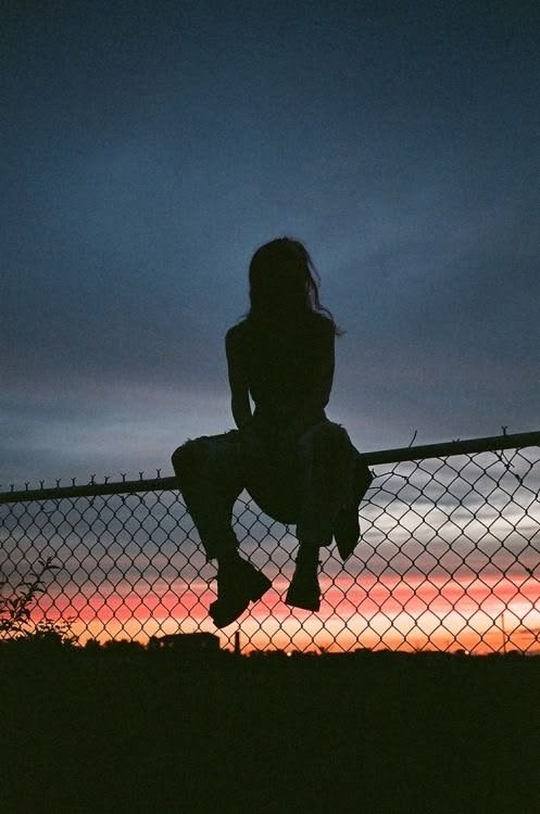 Silhouette d'une adolescente assise en haut d'un grillage, de dos, devant un ciel rouge