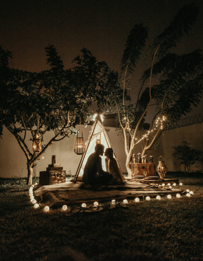 Un homme et une femme face à face devant un tipie éclairé par une guirlande lumineuse entre deux arbres dans la nuit